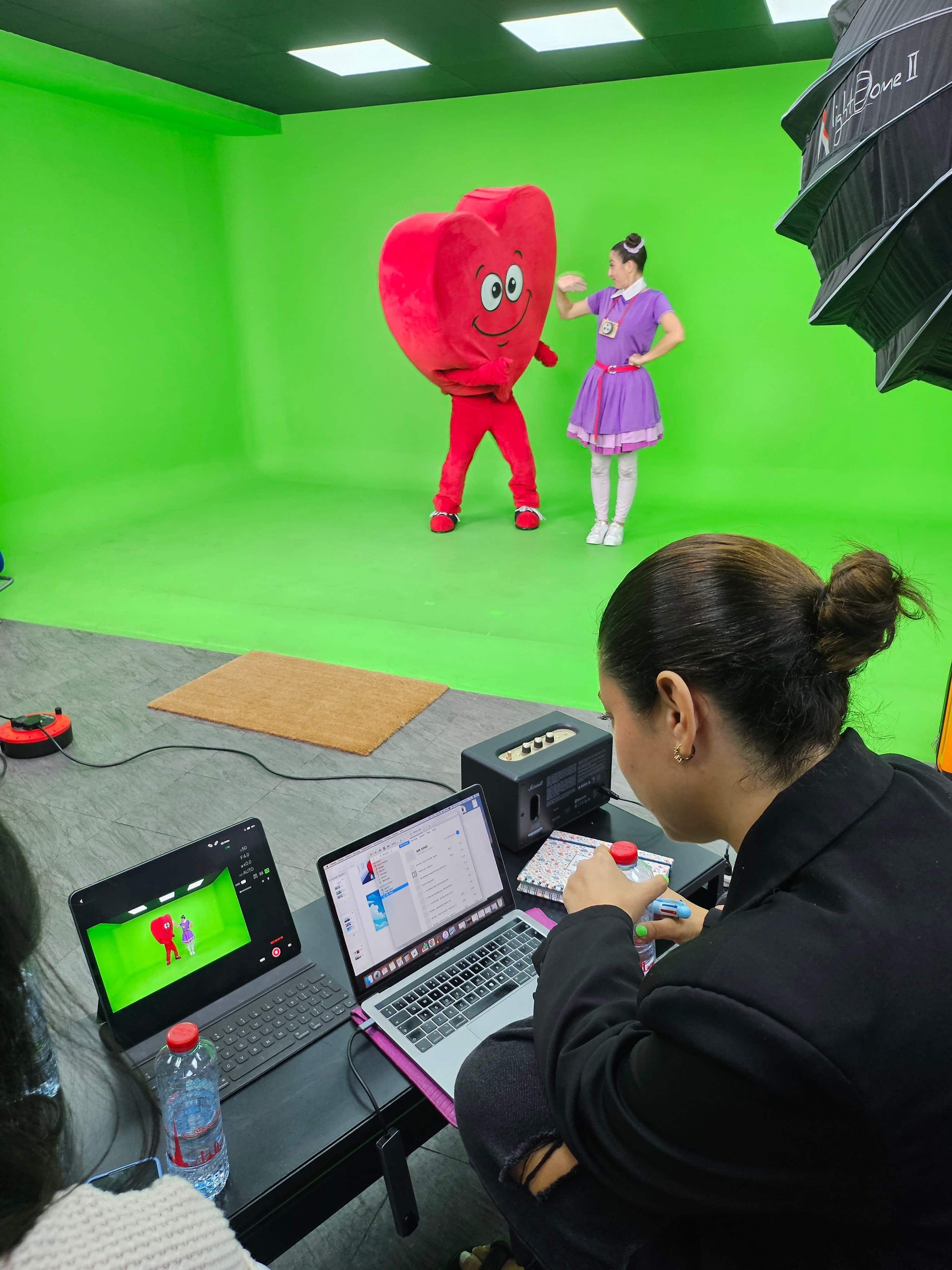 A woman monitors a green screen shoot featuring a dancer and red heart mascot.