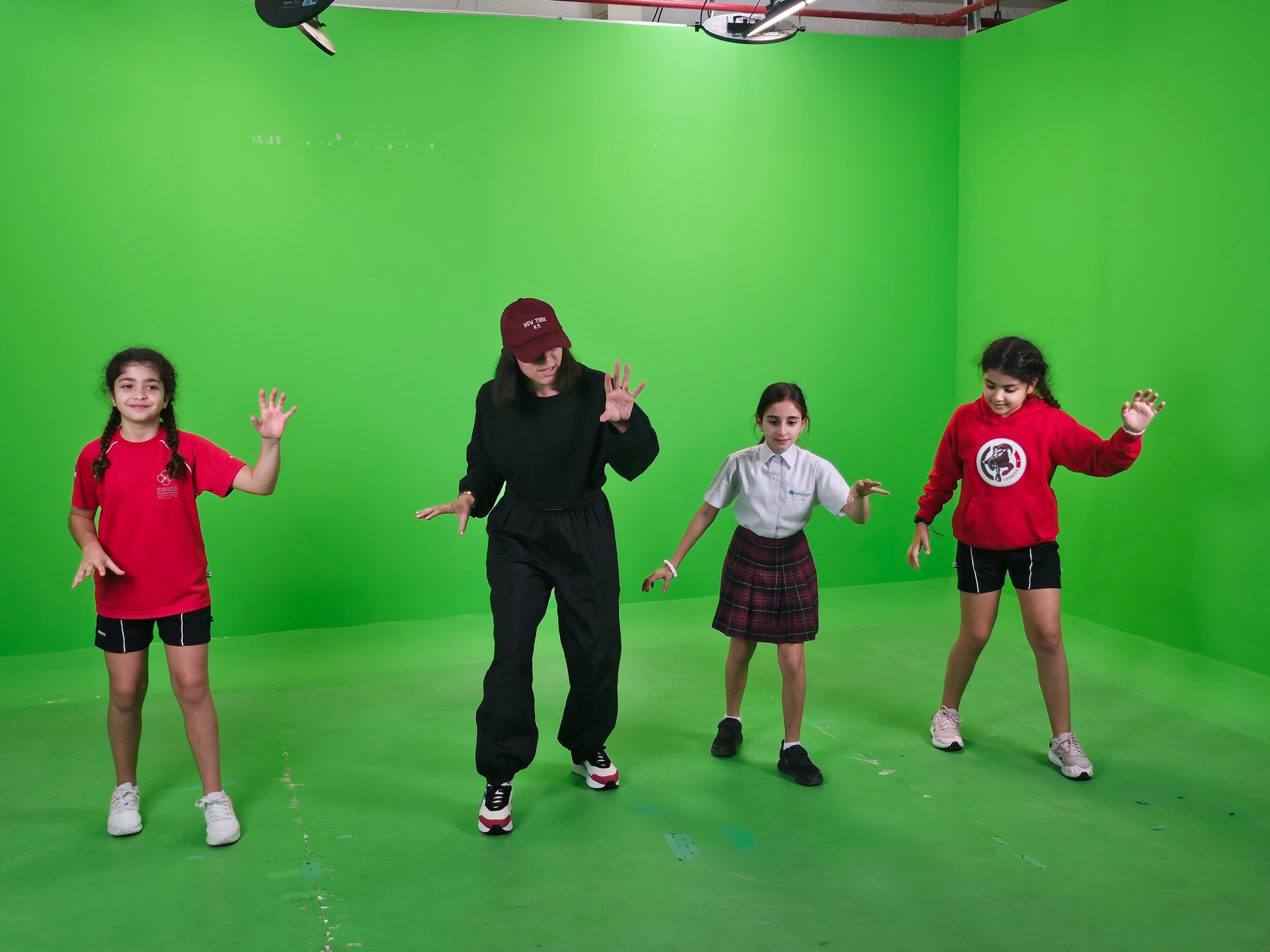 A woman and three girls practice dance moves in a bright green screen studio.