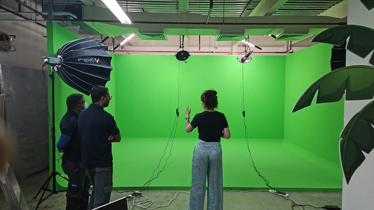 Three people in a professional video studio with a large green screen and lighting equipment.