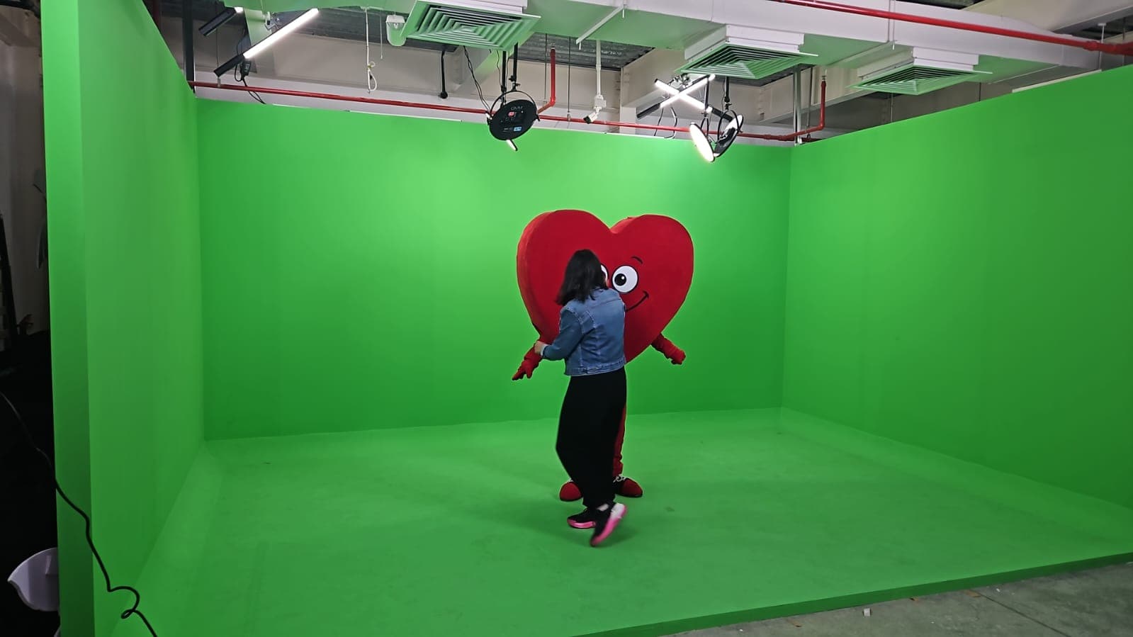 Person in denim jacket interacts with smiling red heart mascot in green screen studio.
